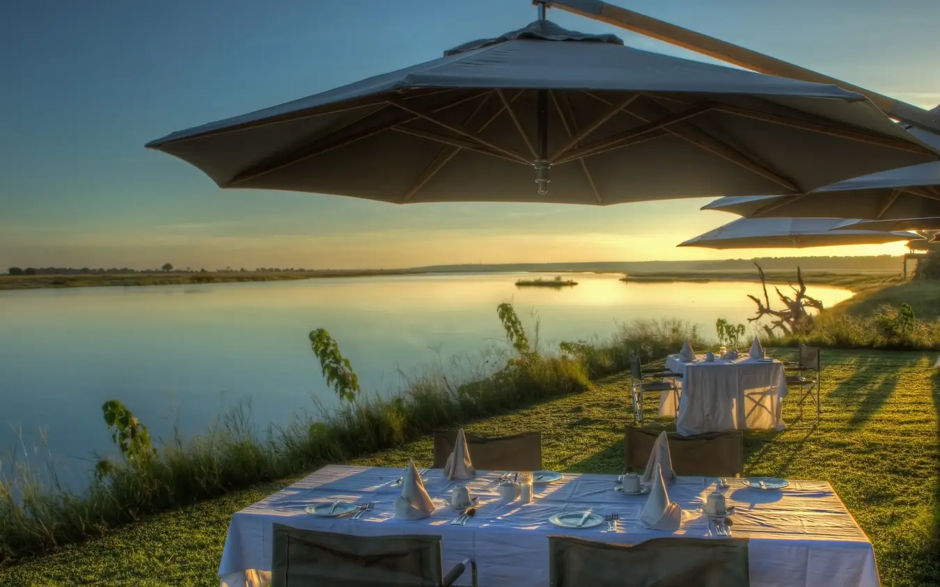 Waterside dining setup with umbrellas overlooking calm river at sunset, Chobe Game Lodge, Chobe, Botswana