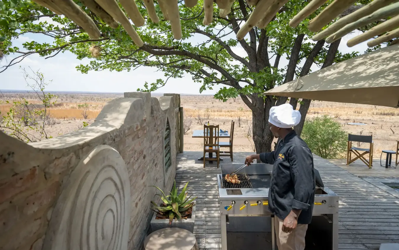Guest grilling meat at outdoor deck under shade tree with savanna vista, Daka Plains Camp, Hwange National Park, Zimbabwe