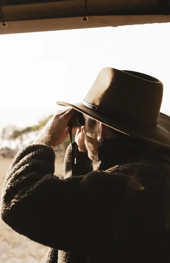 Cowboy adjusting hat while looking through binoculars from safari vehicle, Davison’s Camp, Hwange National Park, Zimbabwe