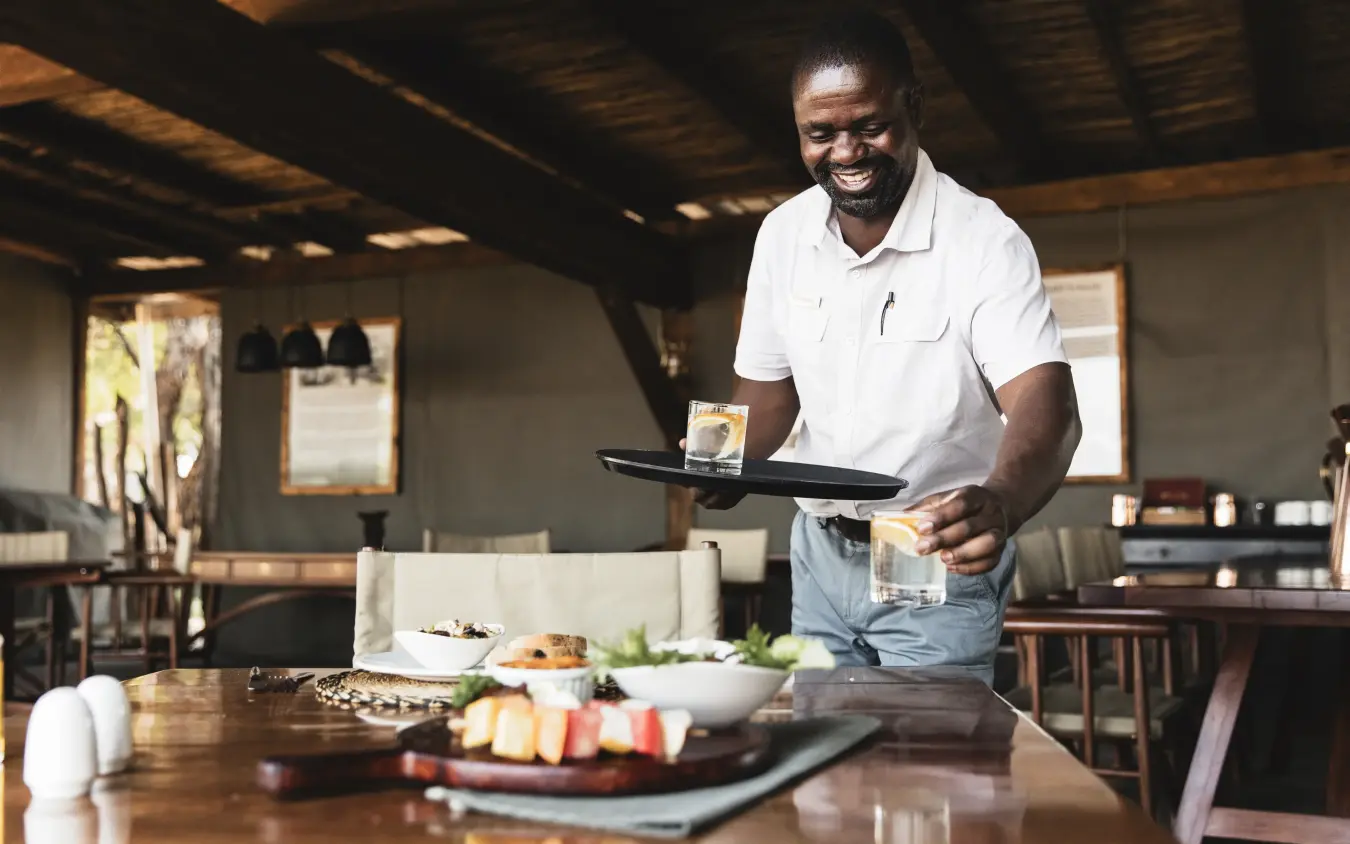 Chef presenting plated dish at wooden dining table in lodge restaurant, Davison’s Camp, Hwange National Park, Zimbabwe