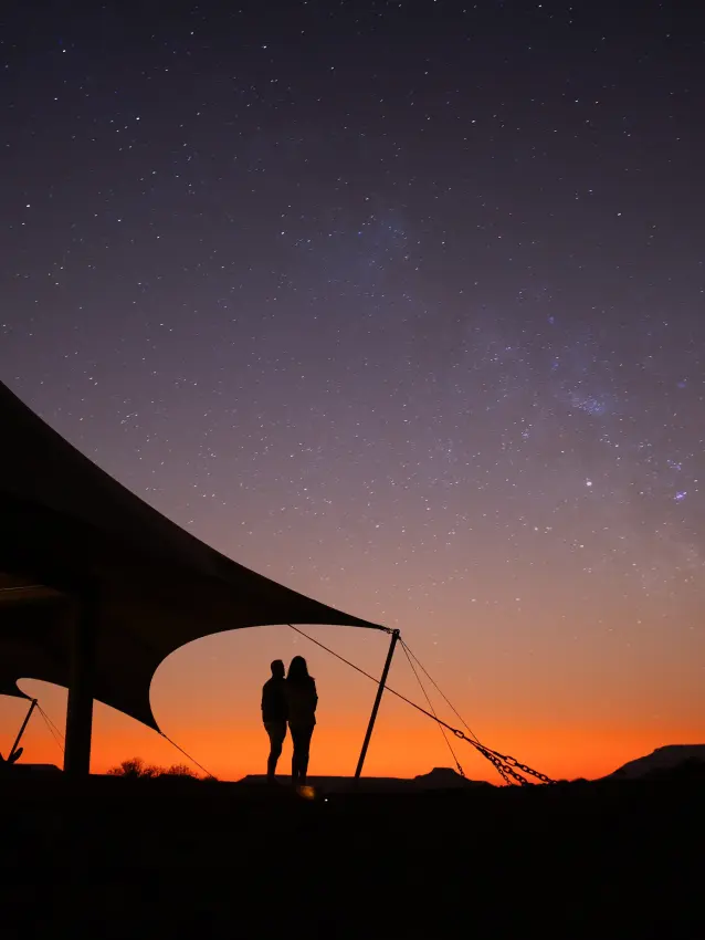 Silhouetted couple standing by tent under starry sky at dusk, Desert Rhino Camp, Damaraland, Namibia