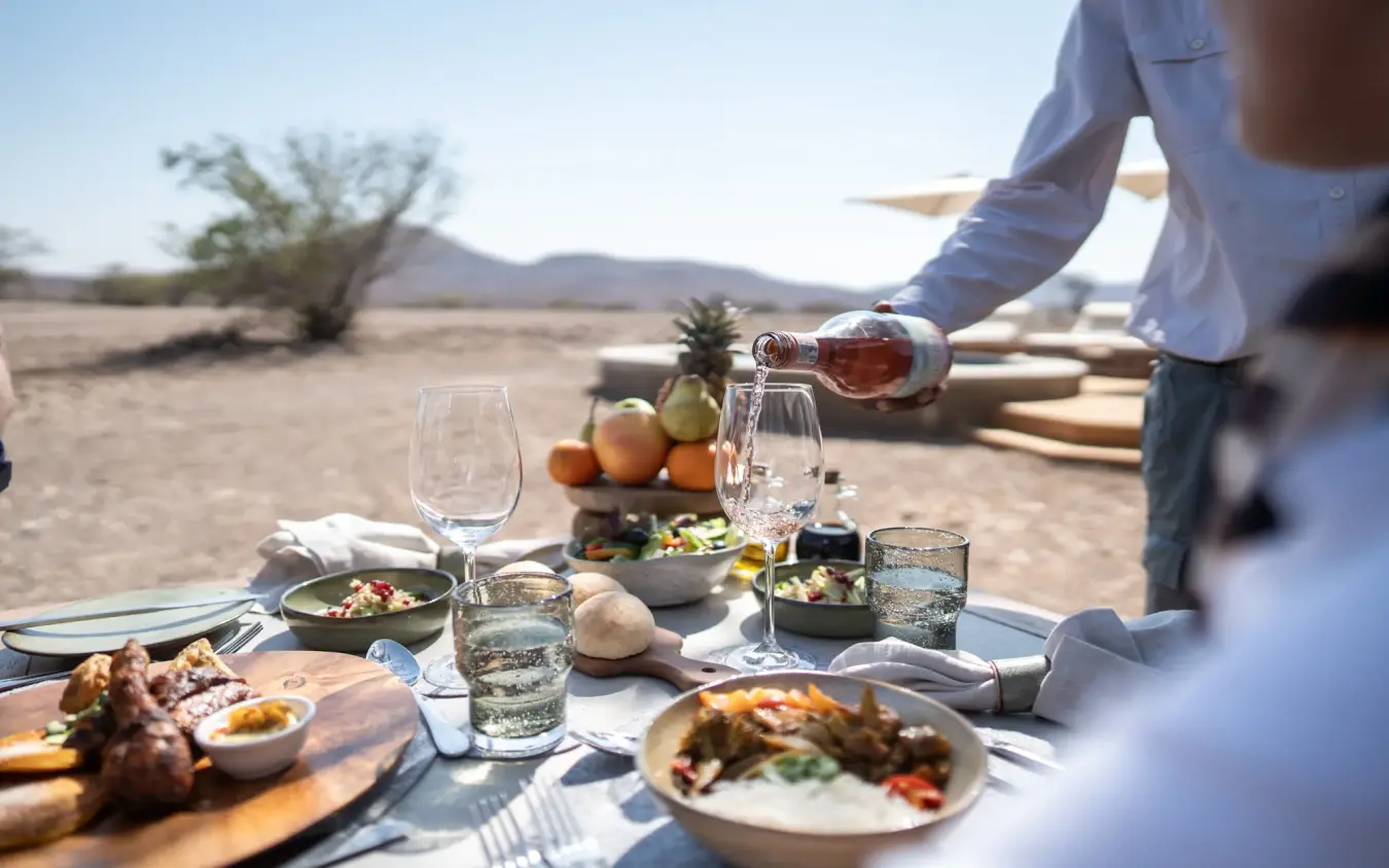 Outdoor dining setup in arid landscape with guests and server pouring wine, Desert Rhino Camp, Damaraland, Namibia