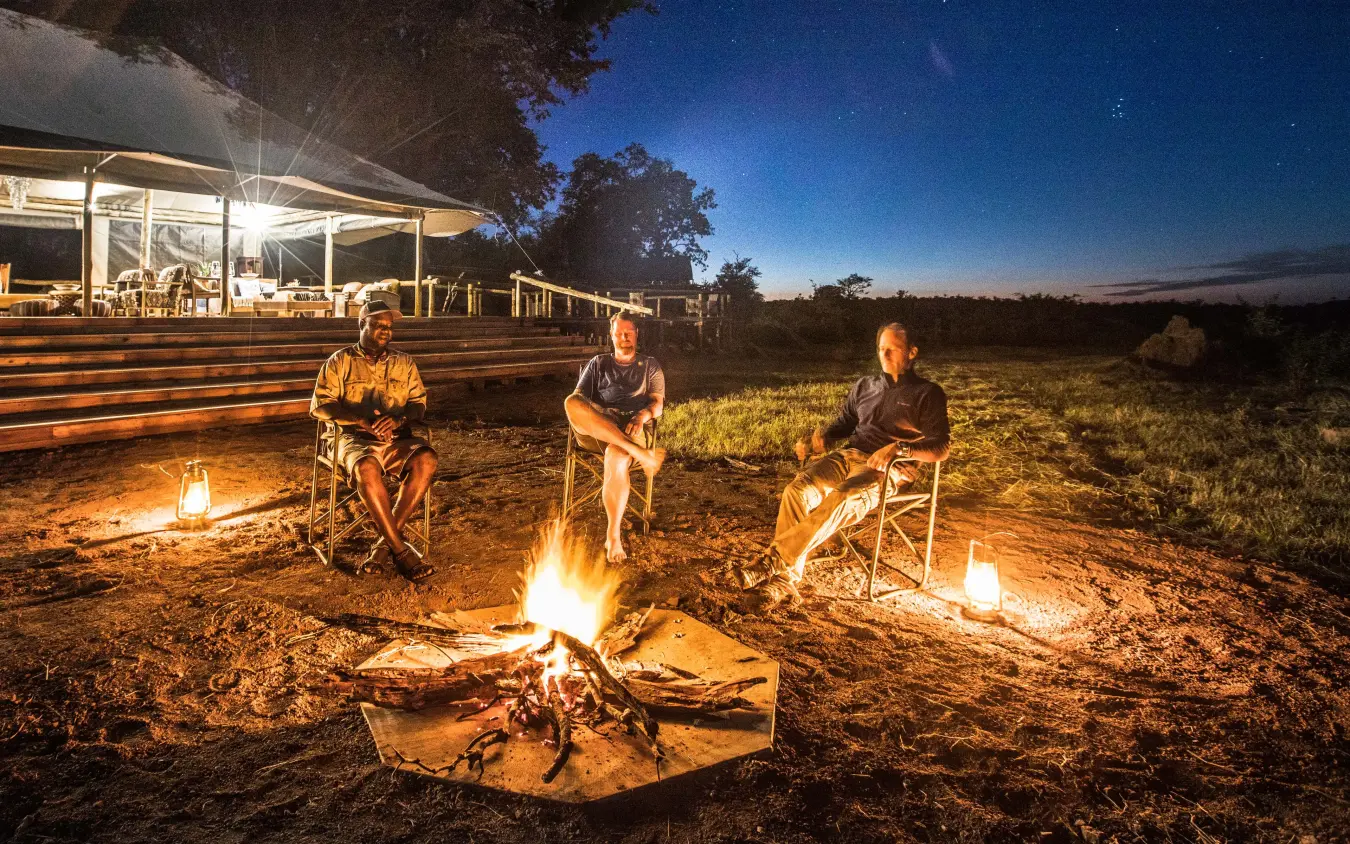 Guests around campfires at dusk with lodge in background, Deteema Springs Camp, Hwange National Park, Zimbabwe