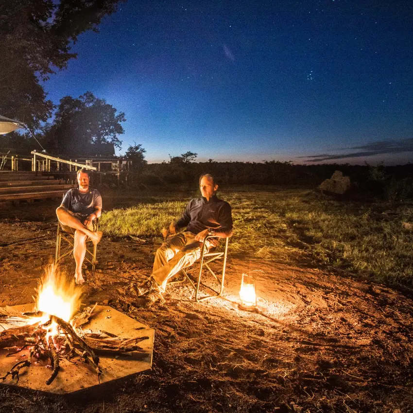 Guests around campfires at dusk with lodge in background, Deteema Springs Camp, Hwange National Park, Zimbabwe