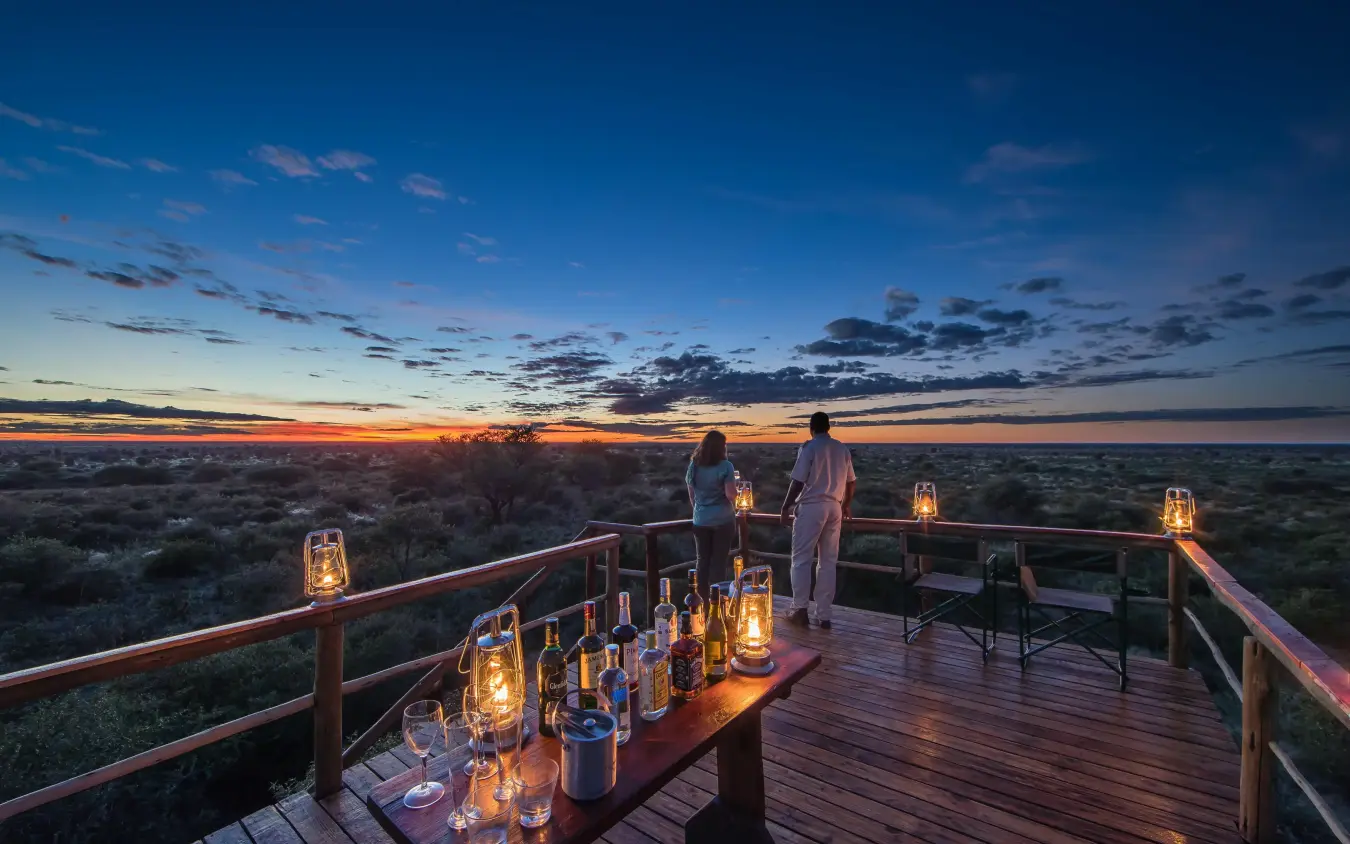Couple enjoying sunset from wooden deck overlooking savanna, Dinaka, Central Kalahari, Botswana