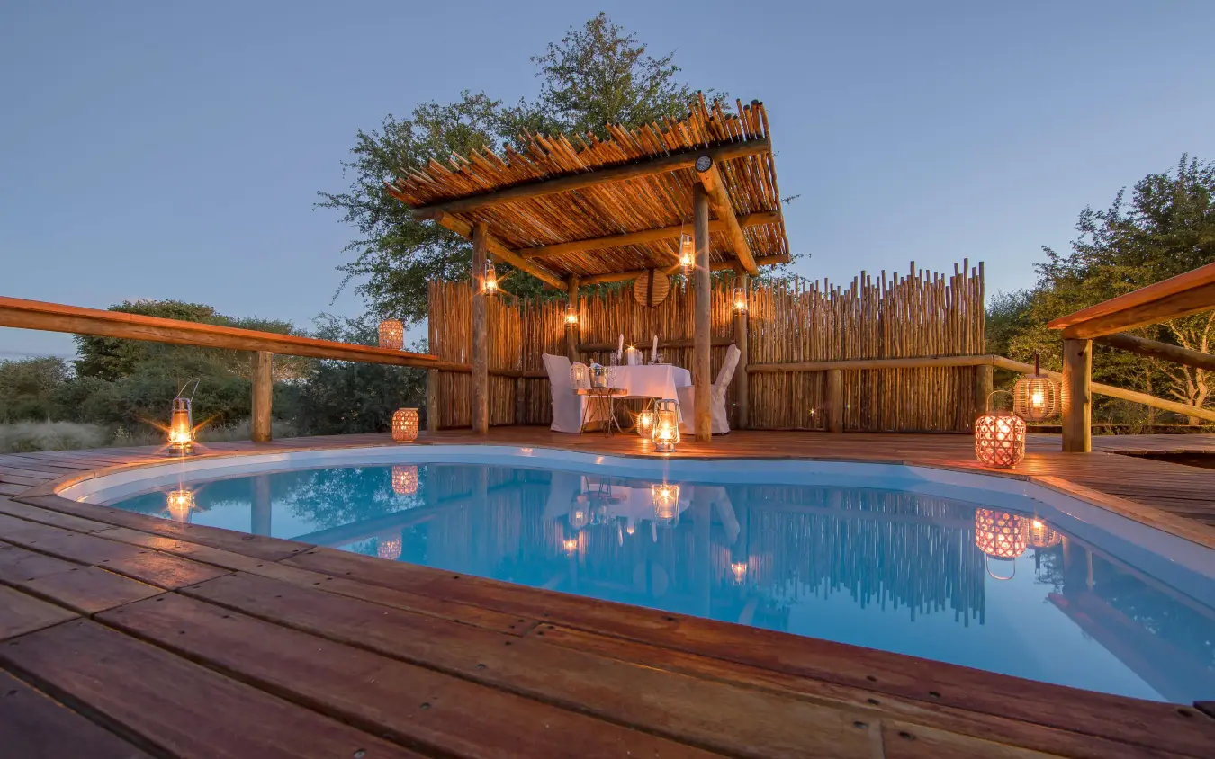 Illuminated resort pool with wooden deck and thatched structures, Dinaka, Central Kalahari, Botswana