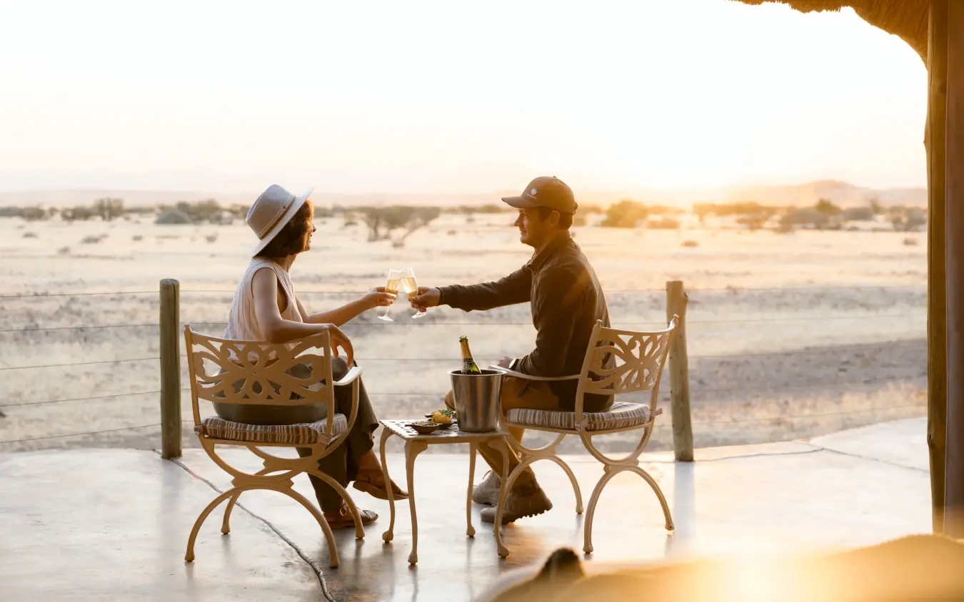 Couple toasting drinks on deck overlooking vast savanna landscape at sunset, Doro Nawas Camp, Damaraland, Namibia