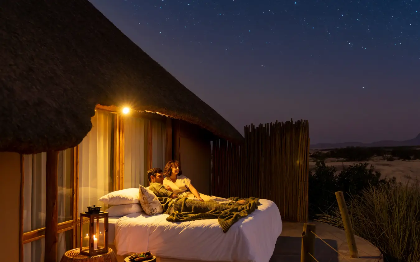 Couple relaxing on bed under stars at open-air safari lodge at night, Doro Nawas Camp, Damaraland, Namibia