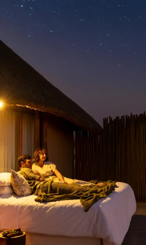 Couple relaxing on bed under stars at open-air safari lodge at night, Doro Nawas Camp, Damaraland, Namibia