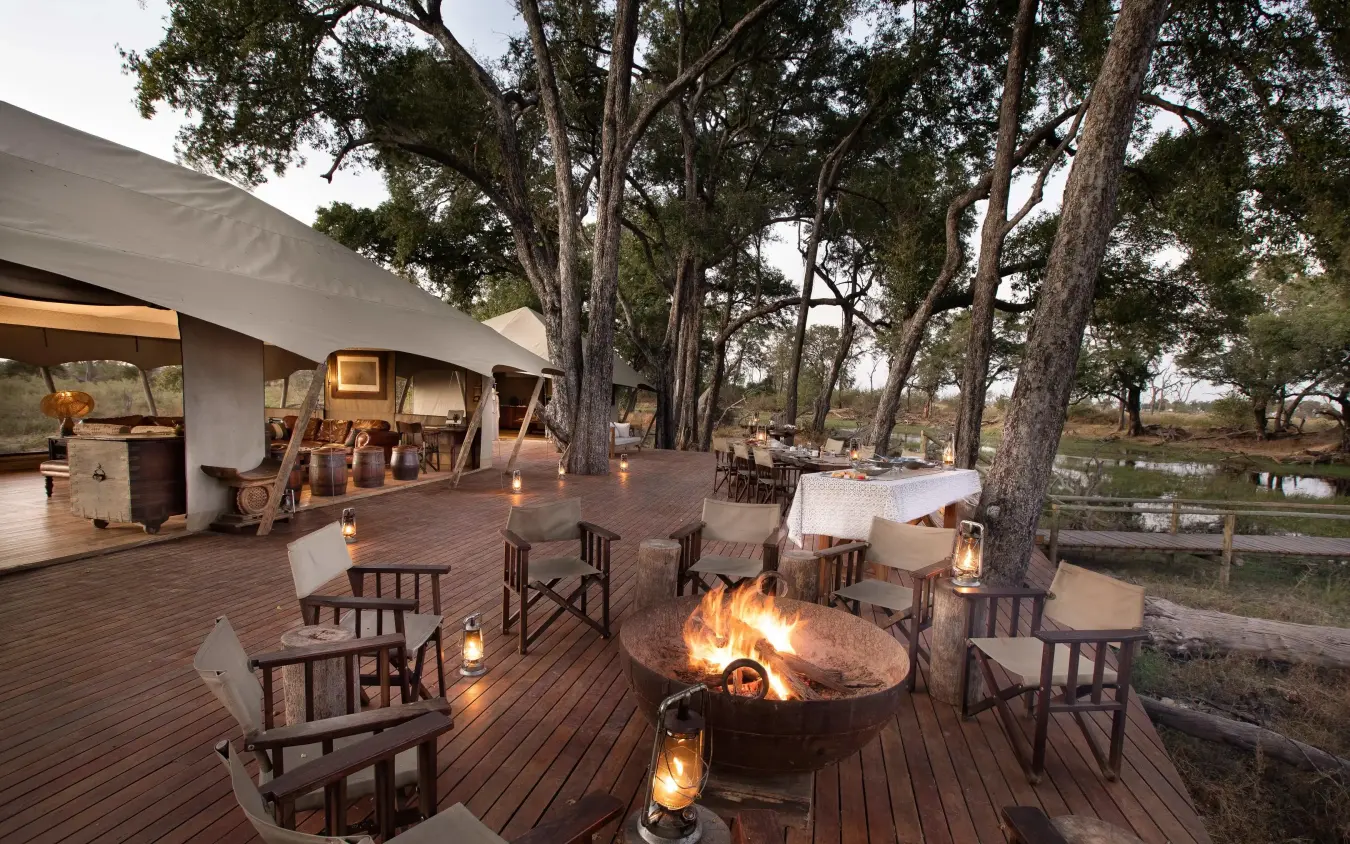 Wooden deck with firepit and seating under large trees, Duba Explorers Camp, The Okavango Delta, Botswana