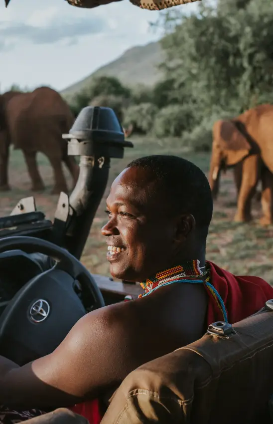 Safari guide smiling while driving open vehicle with elephants in background, Elephant Bedroom Camp, Samburu Reserve, Kenya