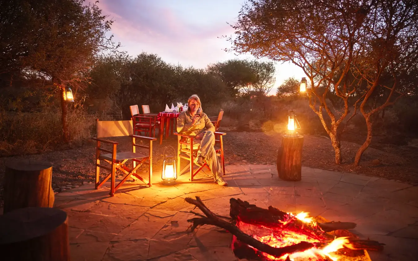 Woman seated by campfire at outdoor dinner setup at dusk, Elephant Gorge Camp, Amboseli and Chyulu Hills, Kenya