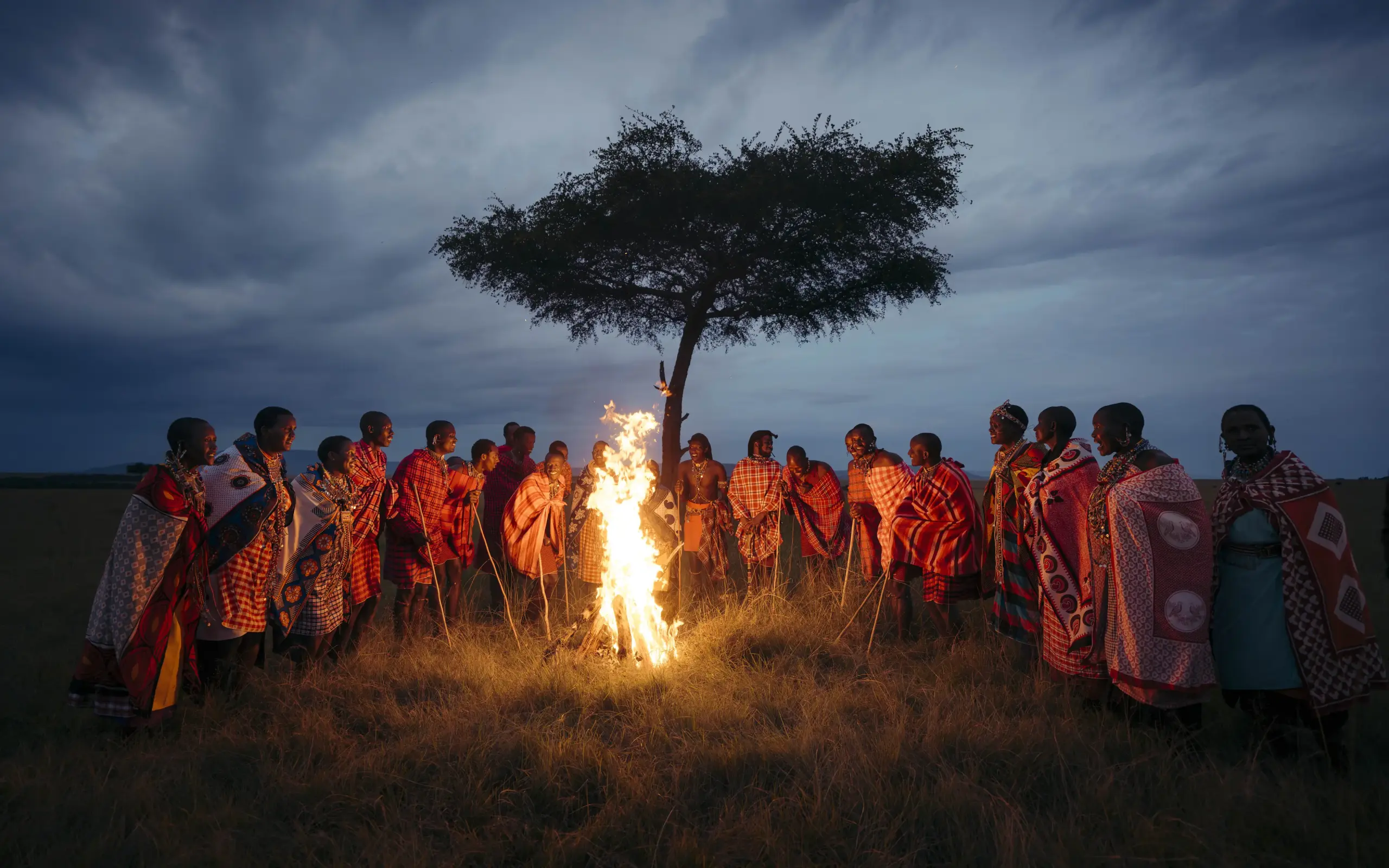 Group gathered around fire under acacia tree at dusk, Emboo Camp, Maasai Mara, Kenya