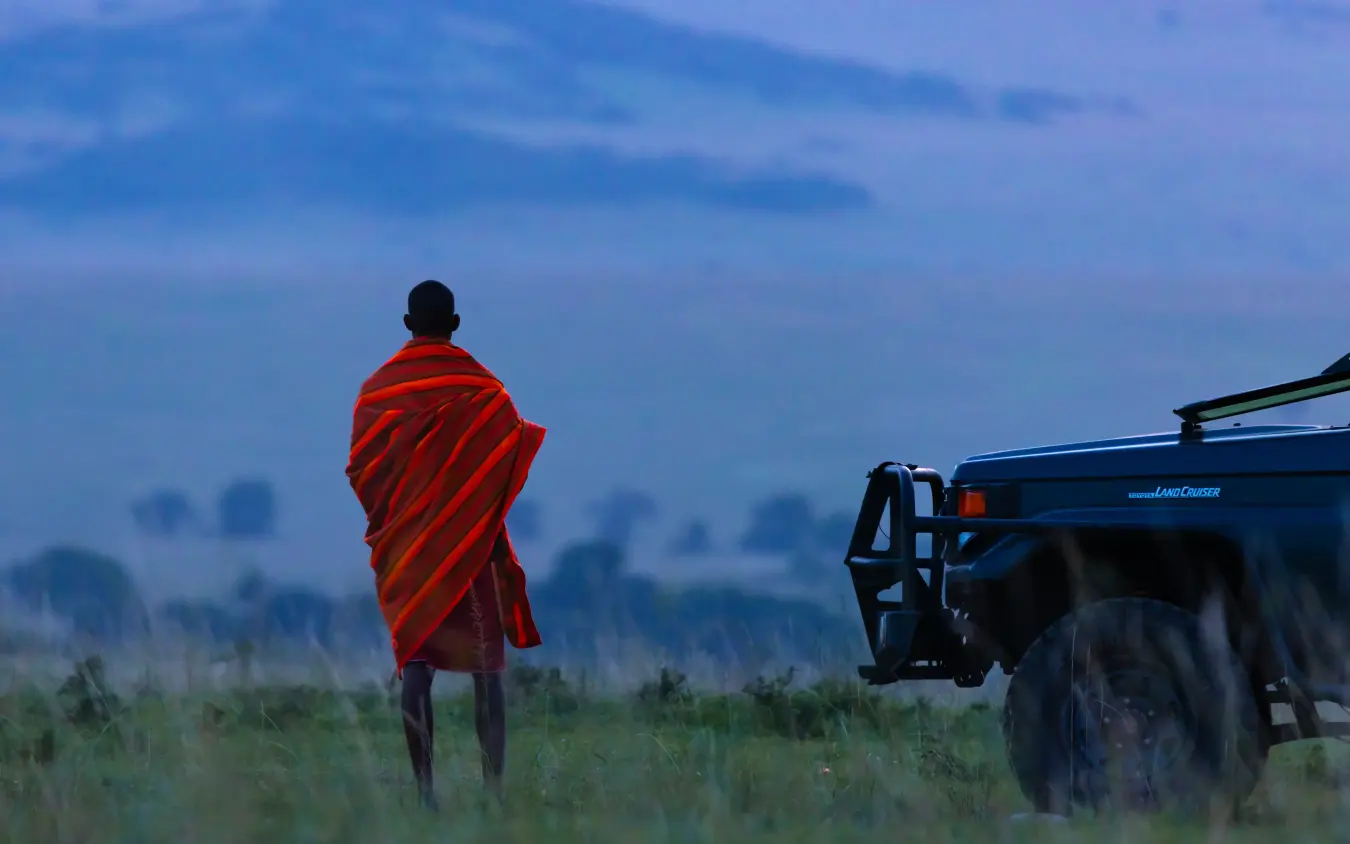 Guide in red shawl standing beside safari vehicle at dawn, Emboo Camp, Maasai Mara, Kenya