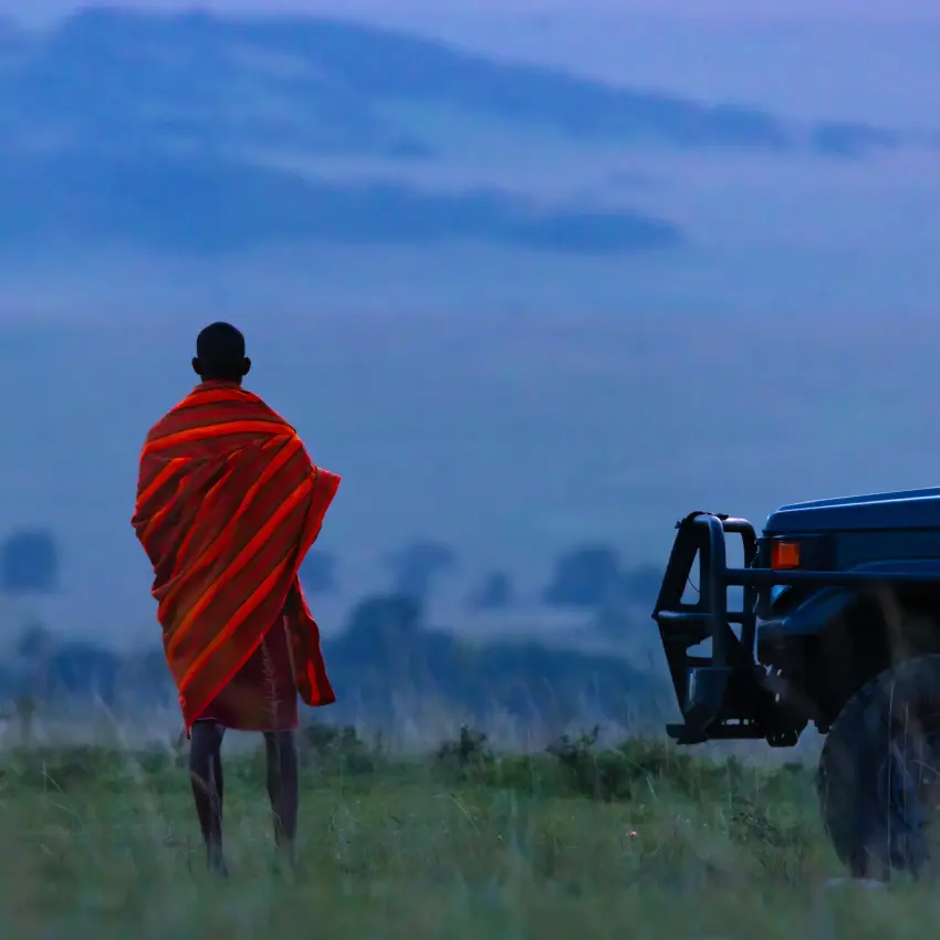 Maasai warrior in red shawl standing near safari vehicle, Maasai Mara, Kenya