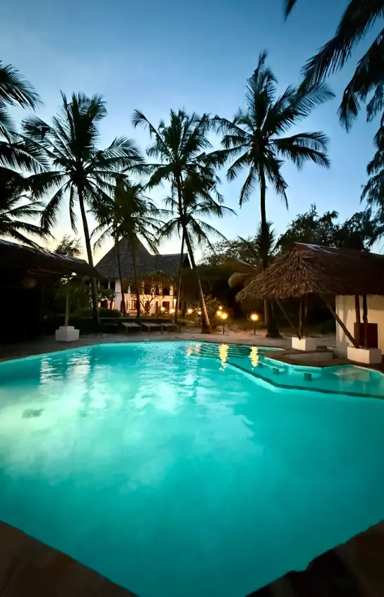 Illuminated swimming pool with palm trees and thatched-roof bungalows at dusk, Enkare Bofa, Kilifi, Kenya