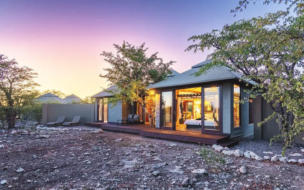 Modern lodge building with illuminated interior at dusk surrounded by trees, Etosha Oberland Lodge, Etosha National Park, Namibia