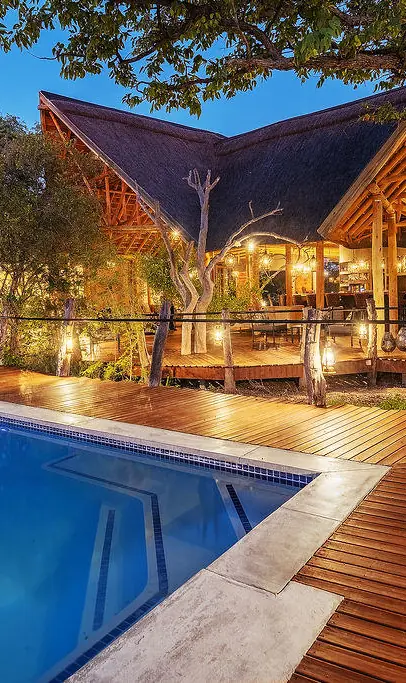 Illuminated swimming pool with wooden deck and thatched lodge building at dusk, Etosha Oberland Lodge, Etosha National Park, Namibia