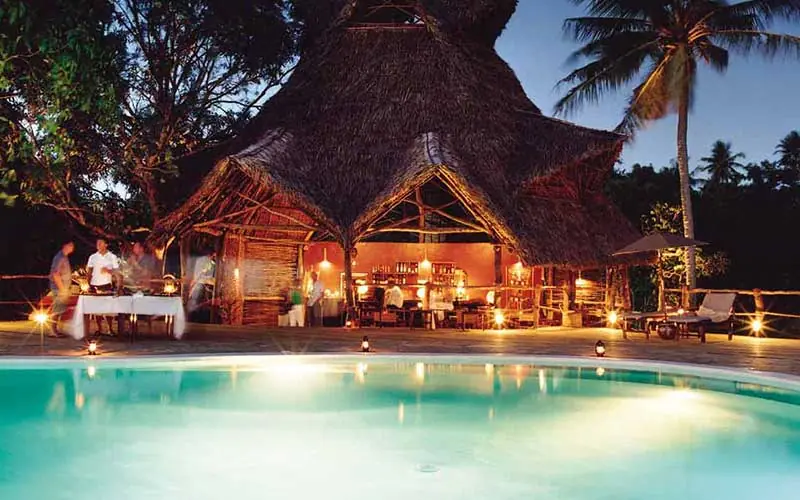 Illuminated thatched lodge buildings beside evening pool, Fundu Lagoon, Zanzibar & Pemba, Tanzania