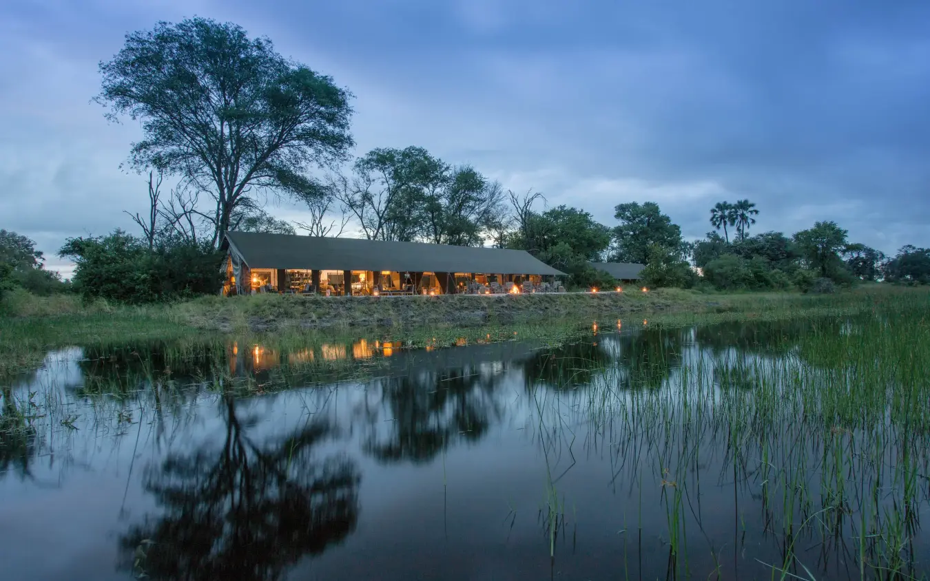 Illuminated main lodge building reflected in still water at dusk, Gomoti Plains, The Okavango Delta, Botswana