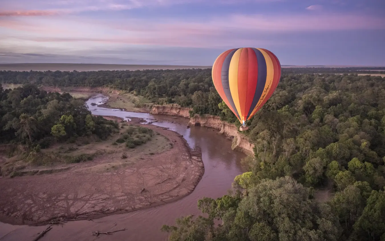 Hot air balloon floating above river and forest at dawn, Governors’ Camp, Maasai Mara, Kenya