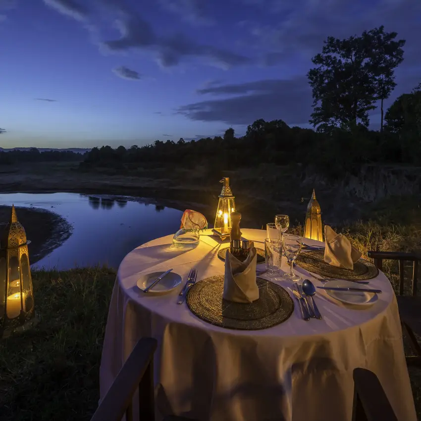 Riverside dinner table set with lanterns at dusk overlooking waterway, Governors’ Camp, Maasai Mara, Kenya