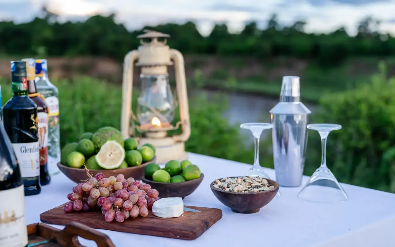Outdoor dining setup with cocktails, fresh fruit and appetizers overlooking wetland landscape, Governors’ Camp, Maasai Mara, Kenya