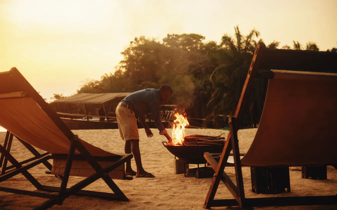 Guest tending to campfire at sunset on sandy beach, Greystoke Mahale, Mahale Mountains, Tanzania