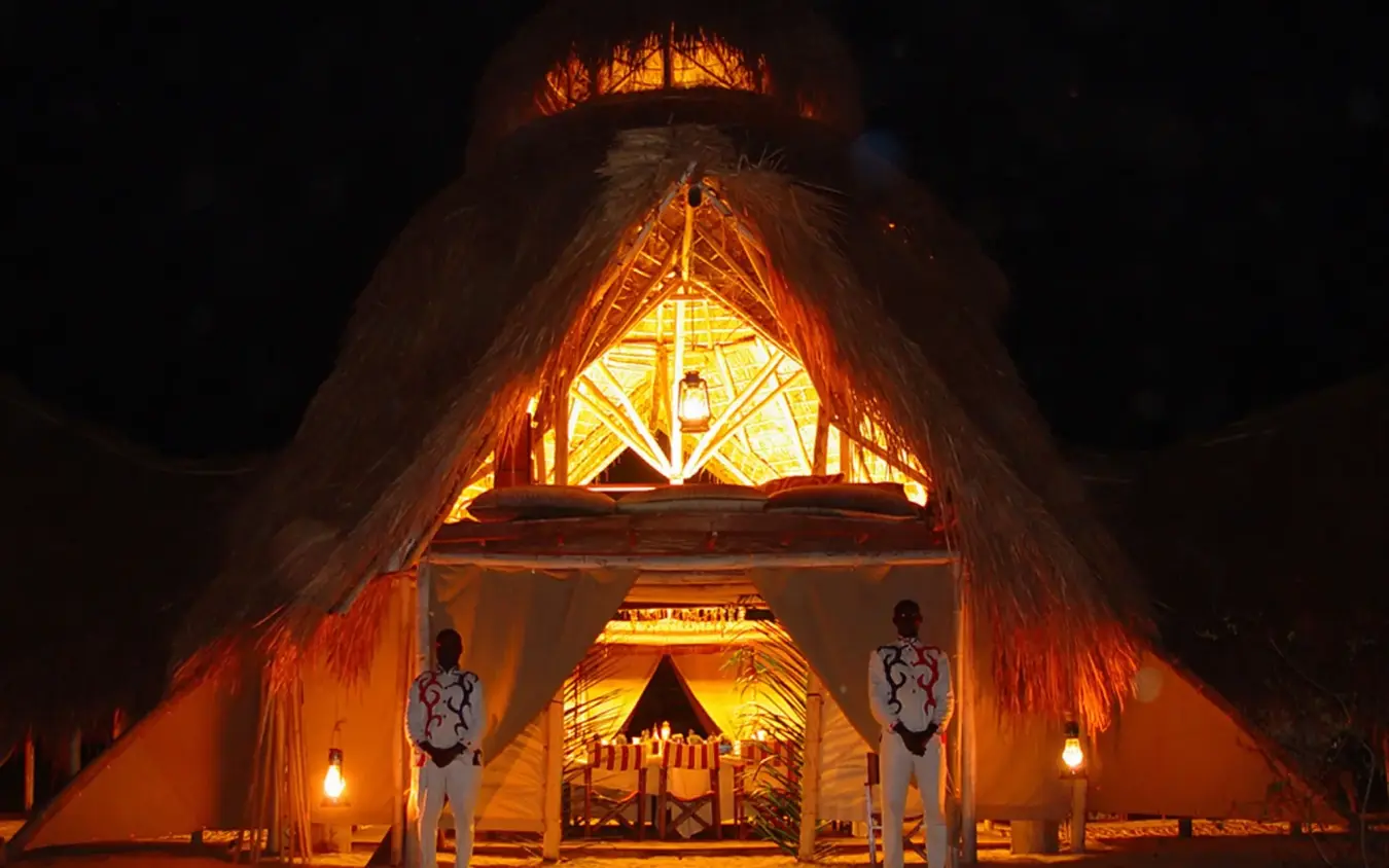 Illuminated thatched-roof lodge entrance at night with staff, Greystoke Mahale, Mahale Mountains, Tanzania