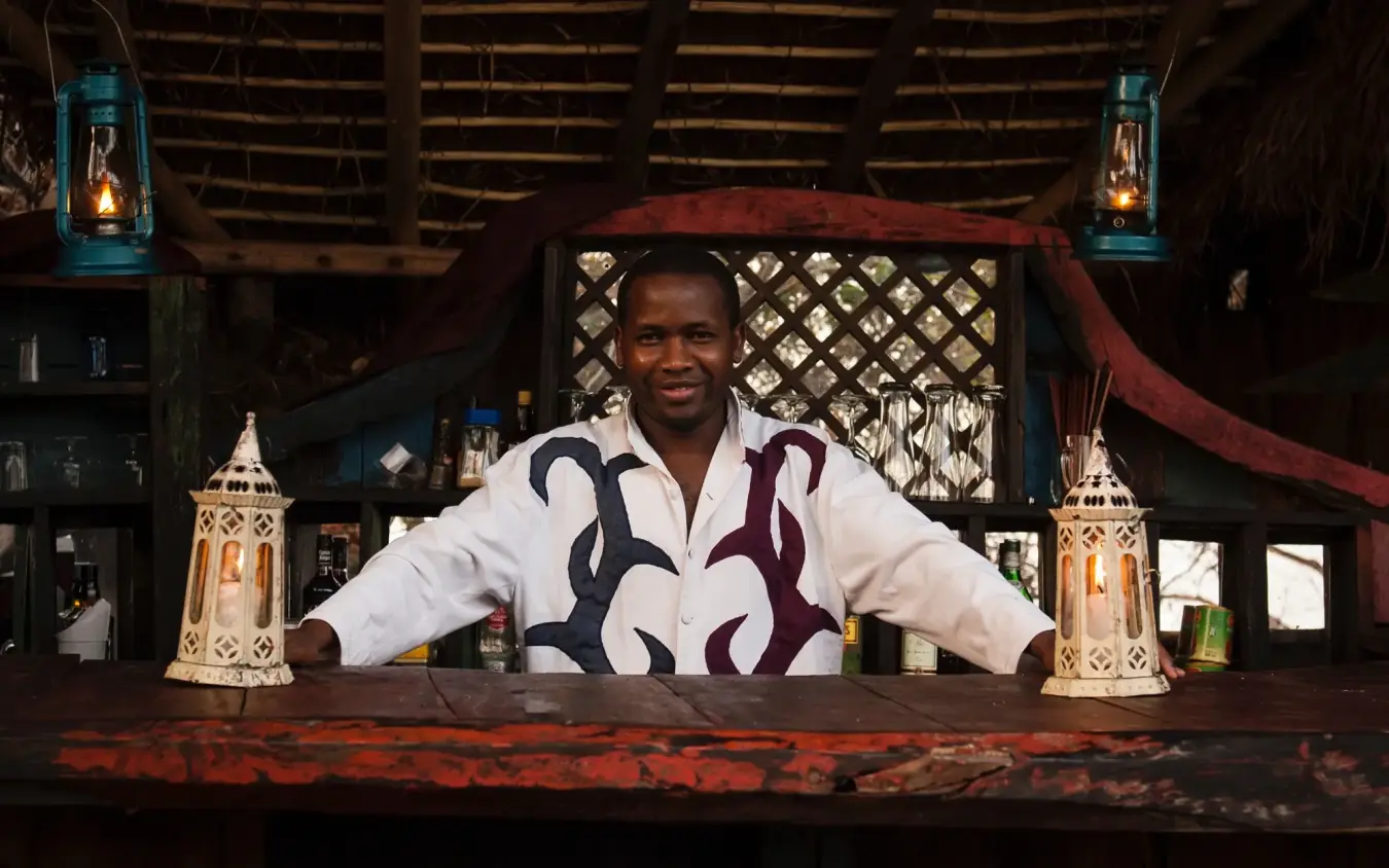 Smiling bartender standing behind wooden lodge bar counter, Greystoke Mahale, Mahale Mountains, Tanzania