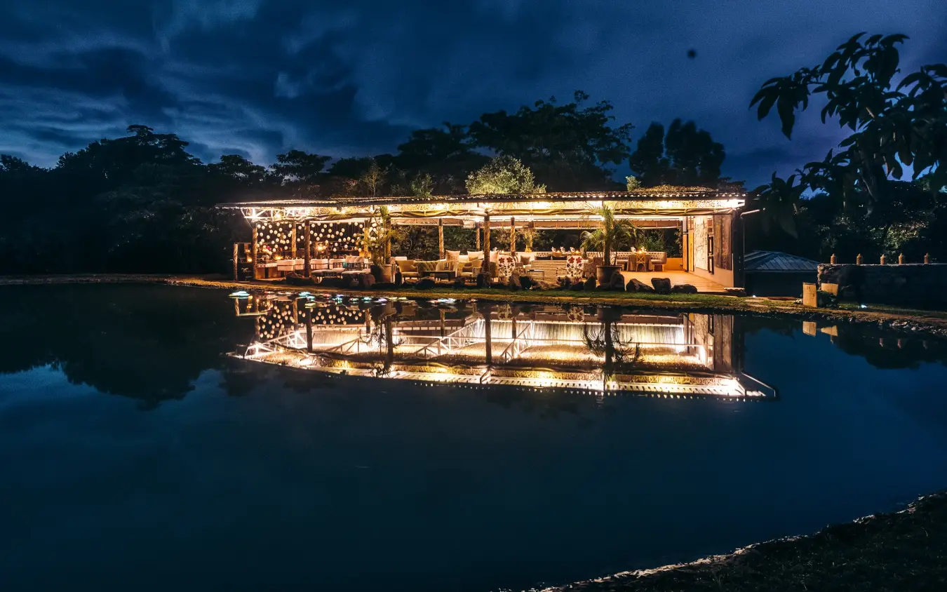 Illuminated waterfront lodge reflecting in calm water at dusk, Hemingways Eden Residence, Nairobi, Kenya