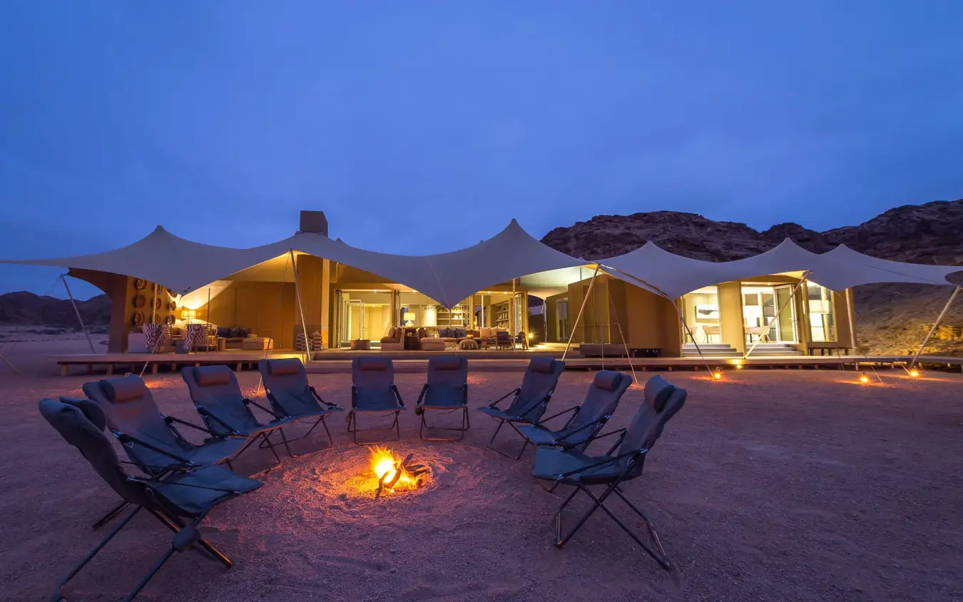 Desert camp with illuminated canvas tents and firepit at dusk, Hoanib Skeleton Coast Camp, Skeleton Coast, Namibia
