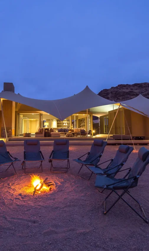 Desert camp with illuminated canvas tents and firepit at dusk, Hoanib Skeleton Coast Camp, Skeleton Coast, Namibia
