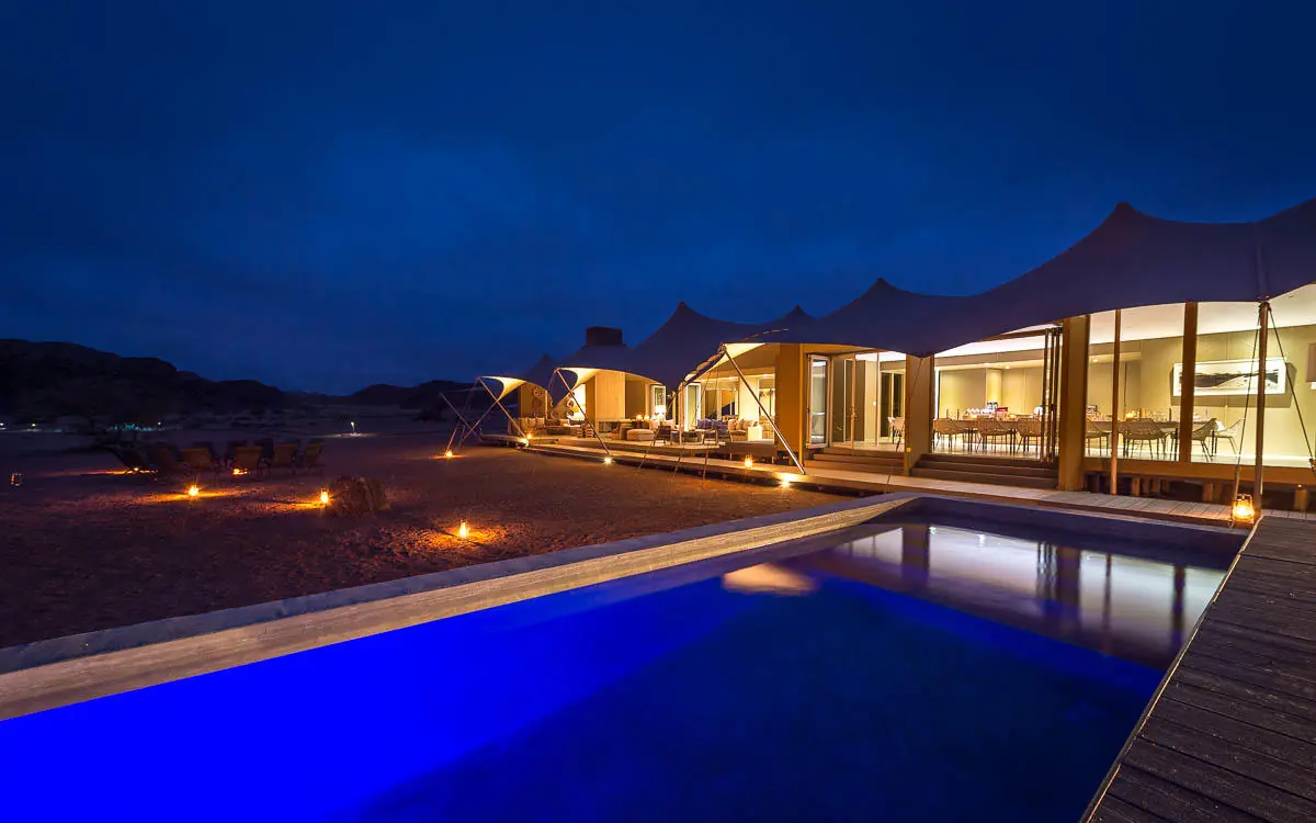 Illuminated swimming pool with desert lodge at dusk, Hoanib Skeleton Coast Camp, Skeleton Coast, Namibia