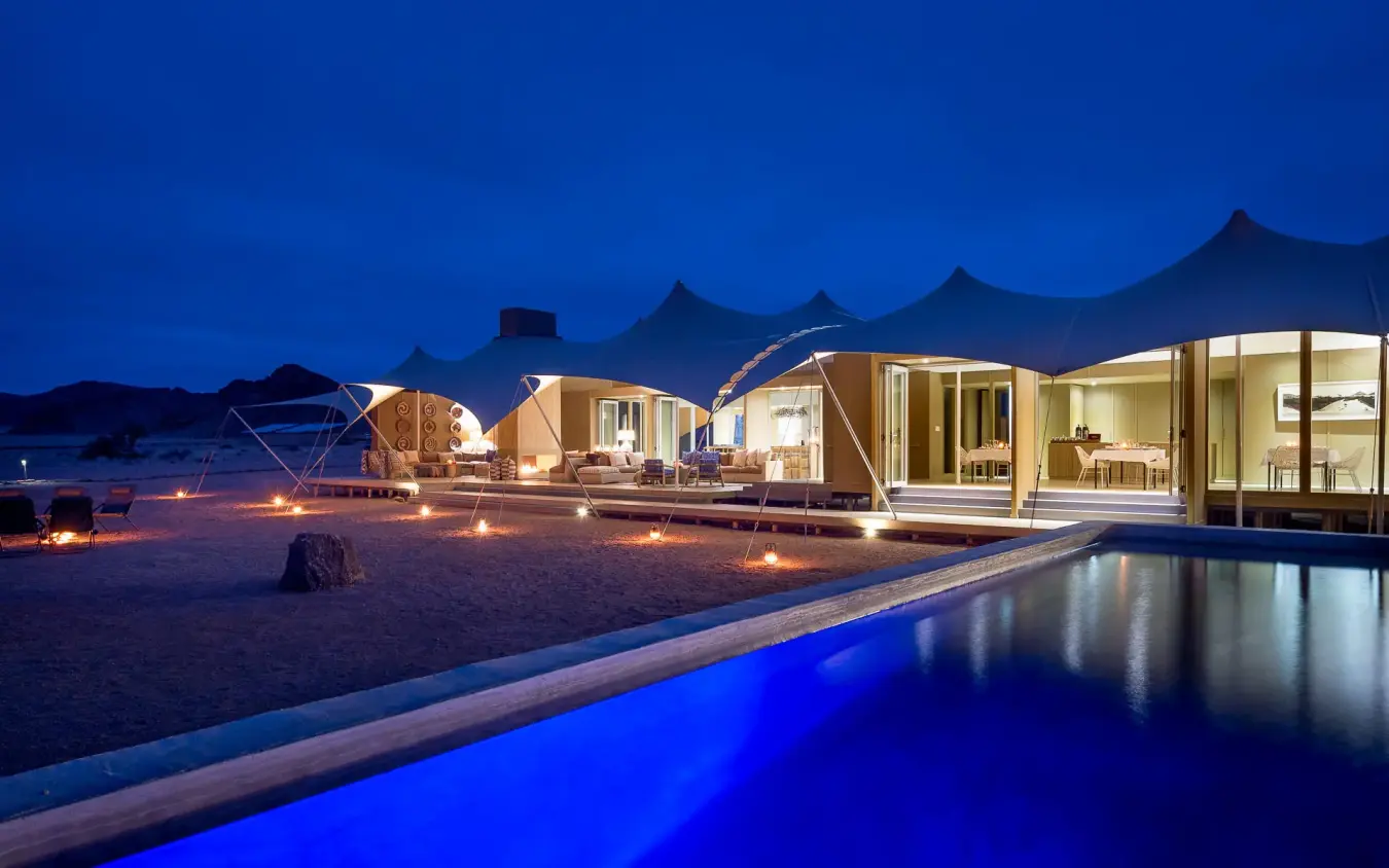 Illuminated pool with tent accommodations and desert landscape at dusk, Hoanib Skeleton Coast Camp, Skeleton Coast, Namibia