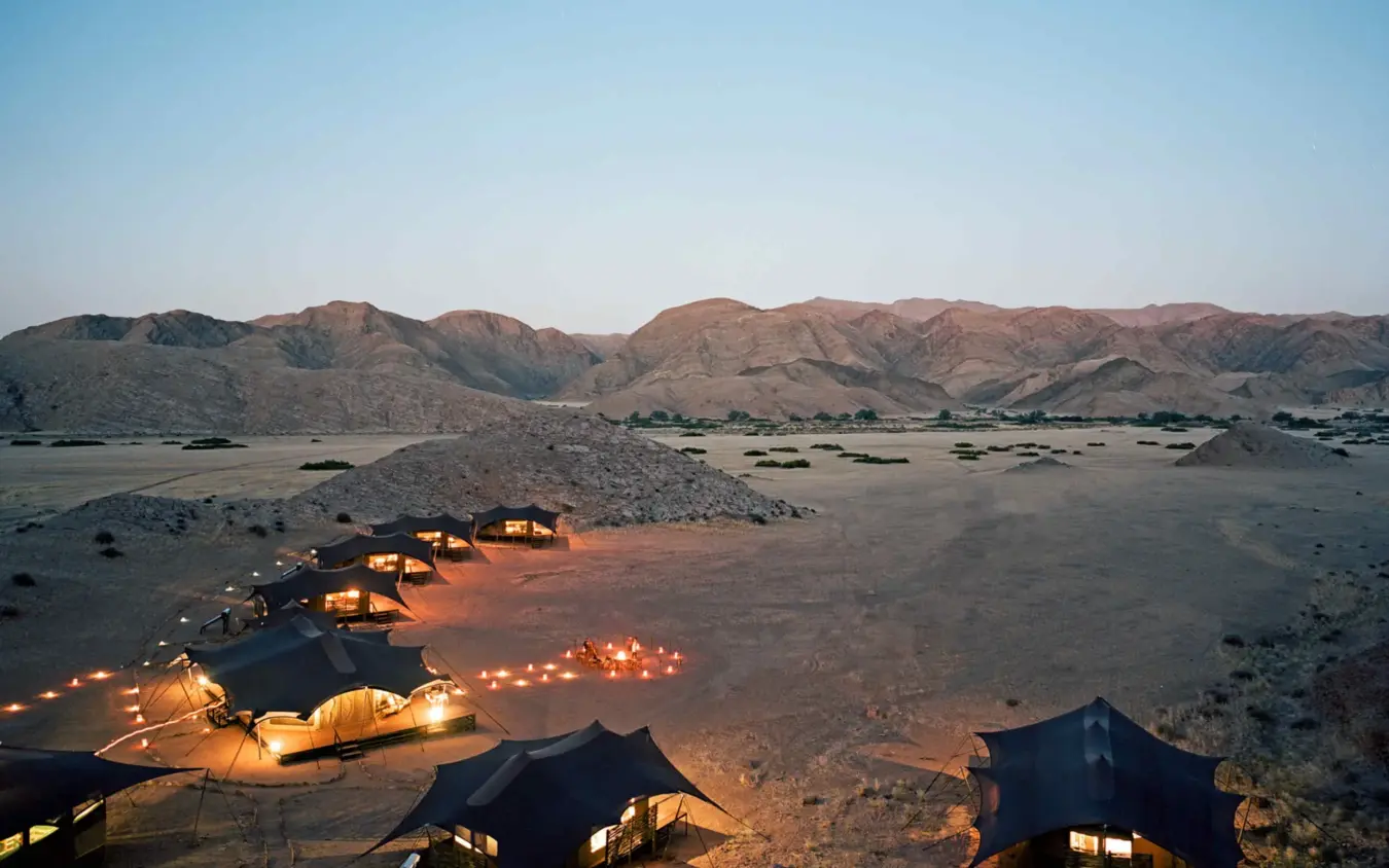 Desert camp with illuminated tents and mountain backdrop at dusk, Hoanib Valley Camp, Skeleton Coast, Namibia