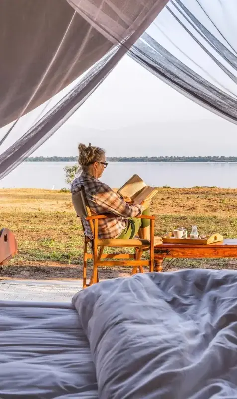 Guest reading in bed with lake view through sheer curtains, Honey Bear Bush Camp, Queen Elizabeth NP, Uganda