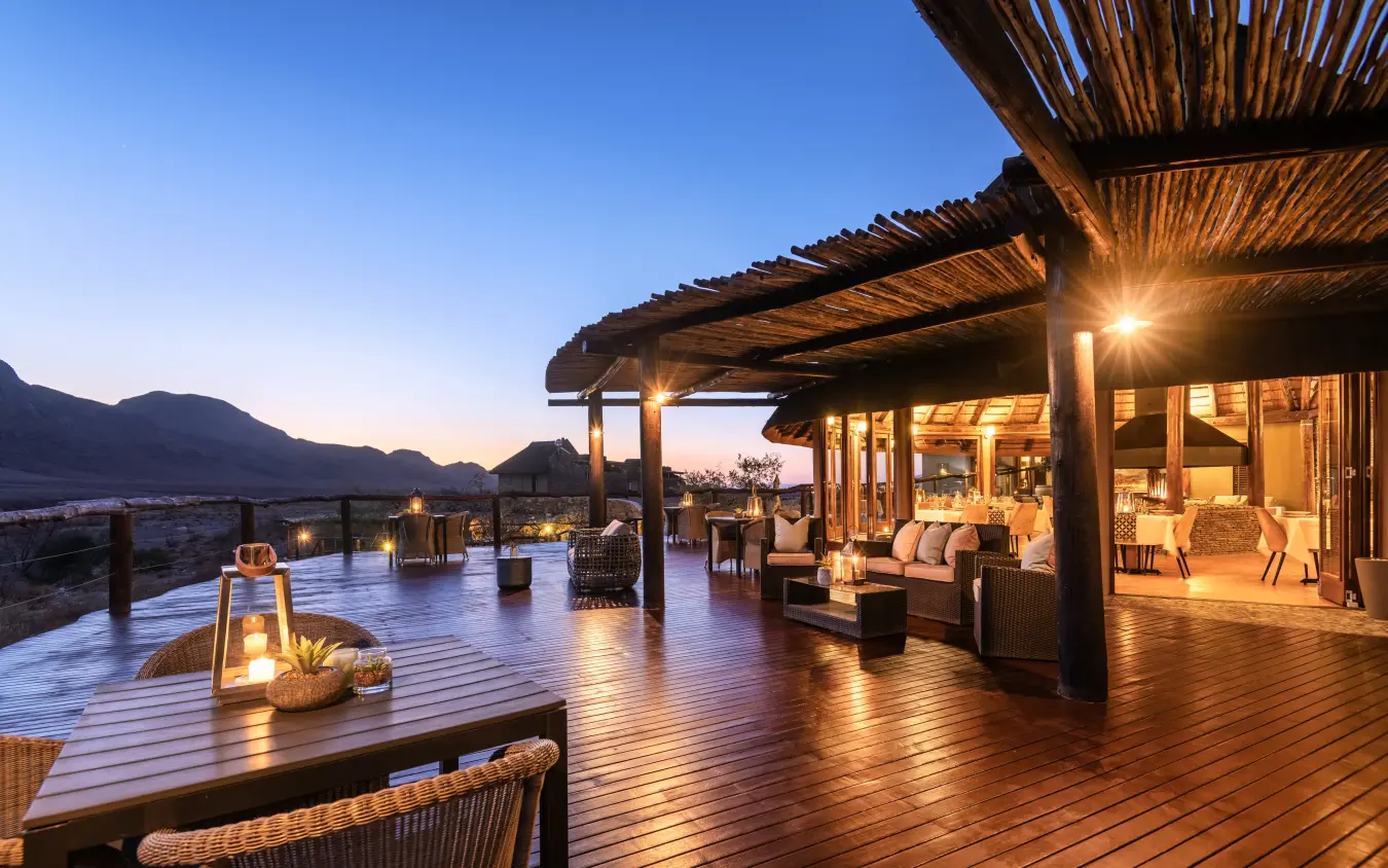 Wooden deck with dining and lounge areas overlooking desert mountains at dusk, Hoodia Desert Lodge, Sossusvlei, Namibia