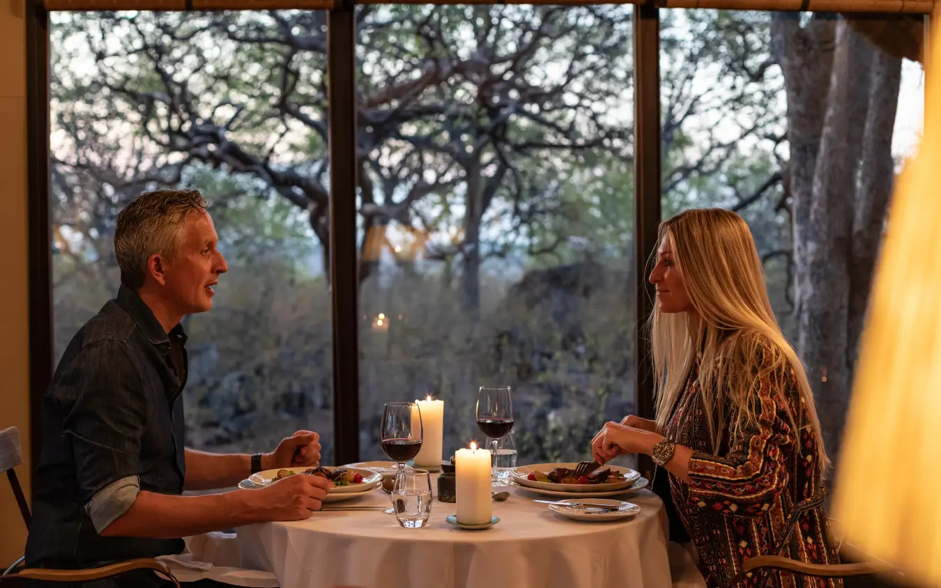 Couple dining at table by large windows overlooking trees at dusk, Horizon by Ongava, Etosha National Park, Namibia