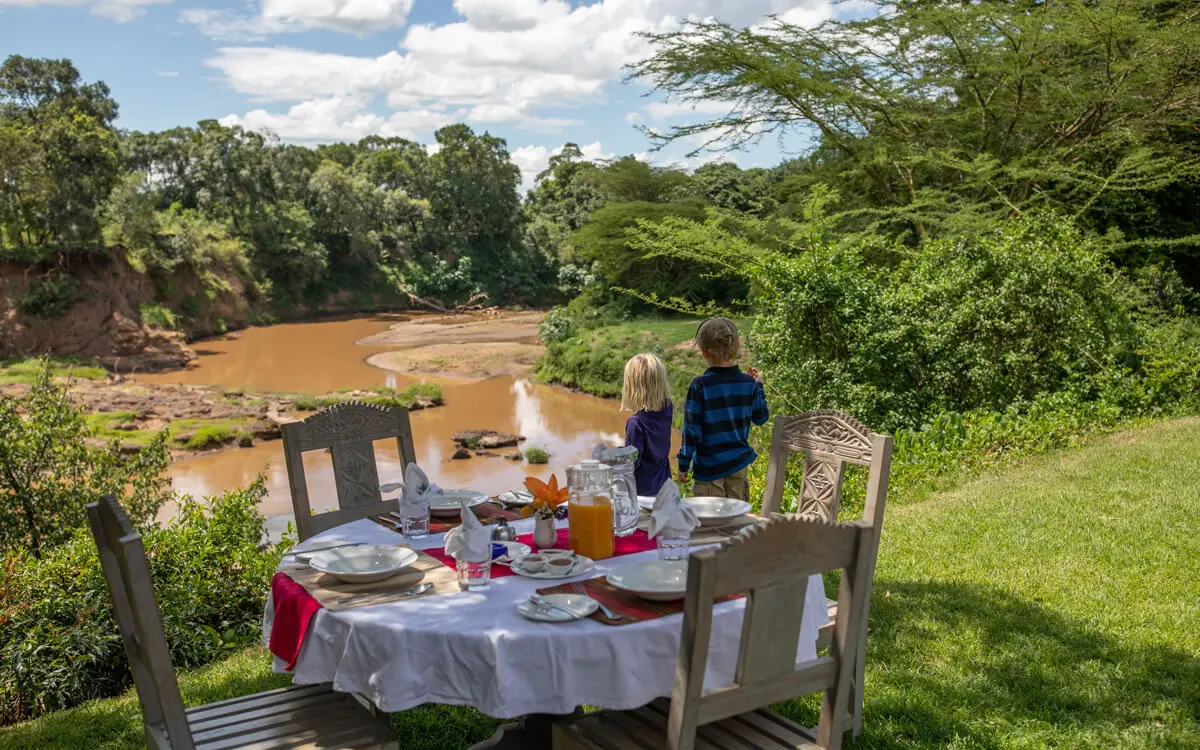 Outdoor riverside dining setup with guests overlooking brown water, House In The Wild, Maasai Mara, Kenya