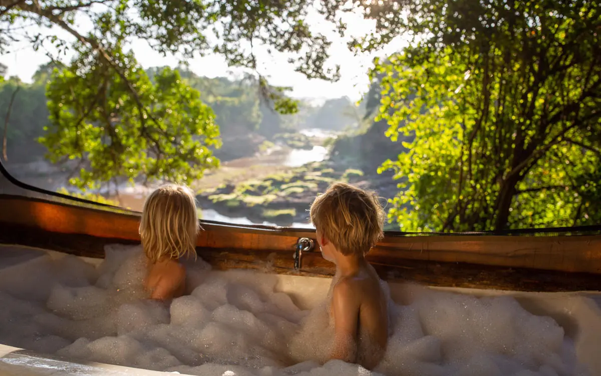 Two children in outdoor bathtub overlooking river landscape, House In The Wild, Maasai Mara, Kenya