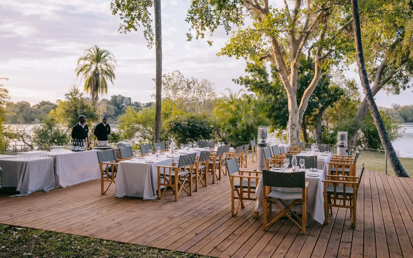 Outdoor dining deck with tables overlooking river and trees at dusk, Ilala Lodge Hotel, Victoria Falls, Zimbabwe