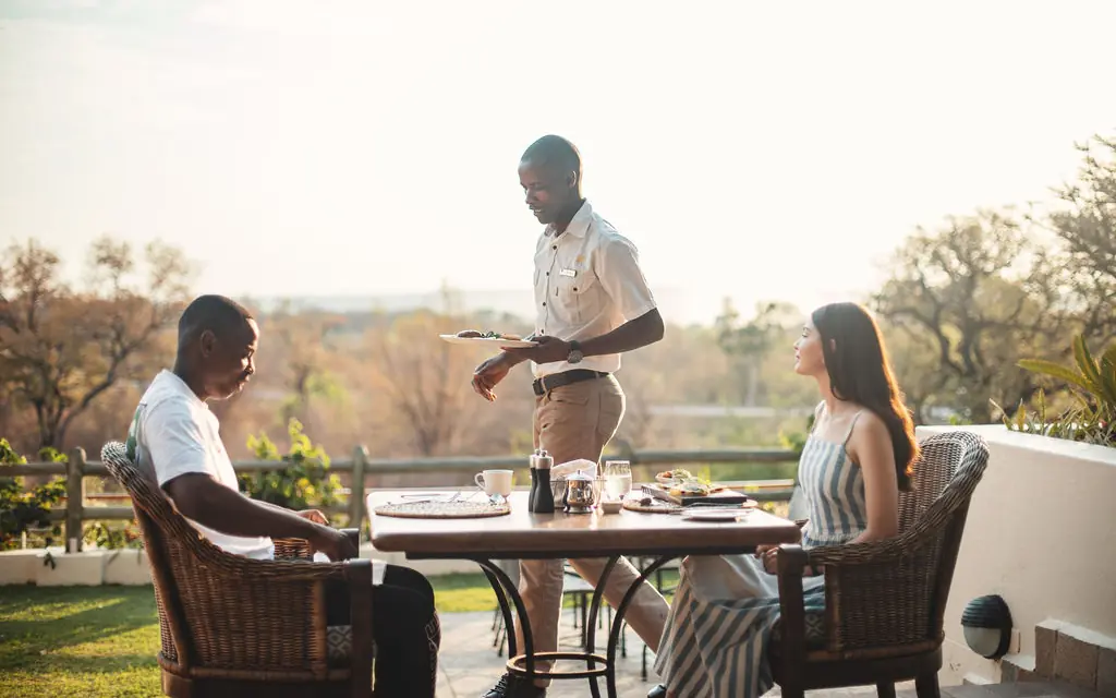 Server presenting meal to guests at outdoor dining table with valley view, Ilala Lodge Hotel, Victoria Falls, Zimbabwe