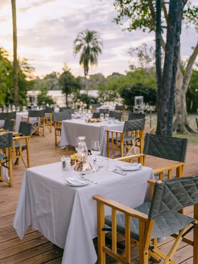 Outdoor dining setup with round tables under trees by riverbank at dusk, Ilala Lodge Hotel, Victoria Falls, Zimbabwe