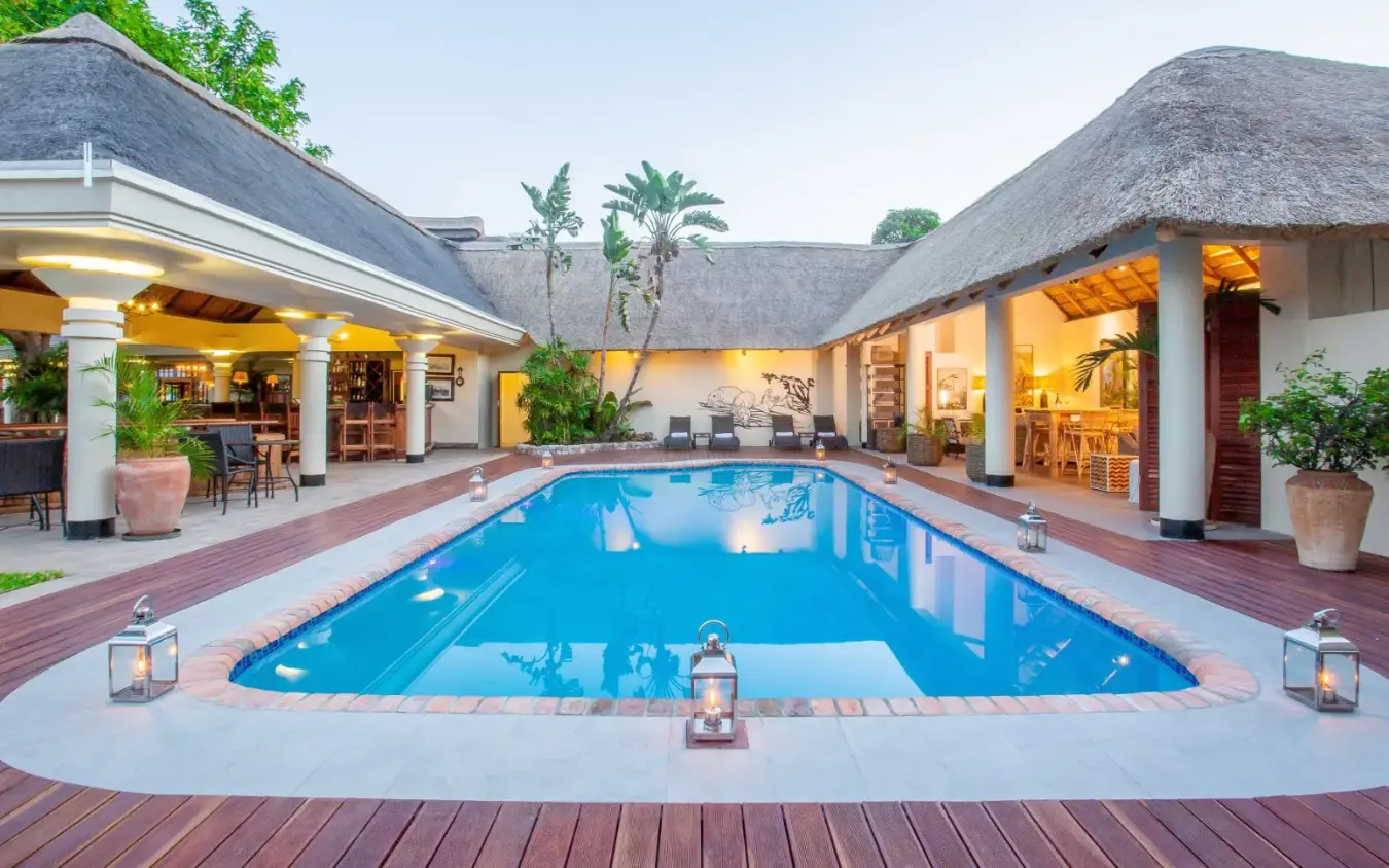 Lit thatch-roof lodge with rectangular pool at dusk, Ilala Lodge Hotel, Victoria Falls, Zimbabwe