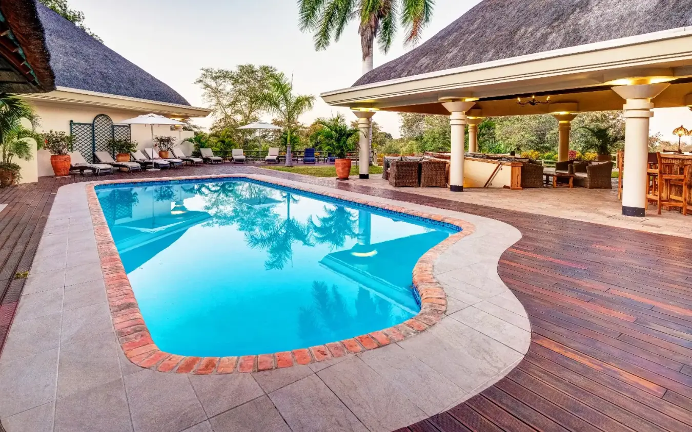 Swimming pool with thatched-roof buildings and wooden deck at sunset, Ilala Lodge Hotel, Victoria Falls, Zimbabwe