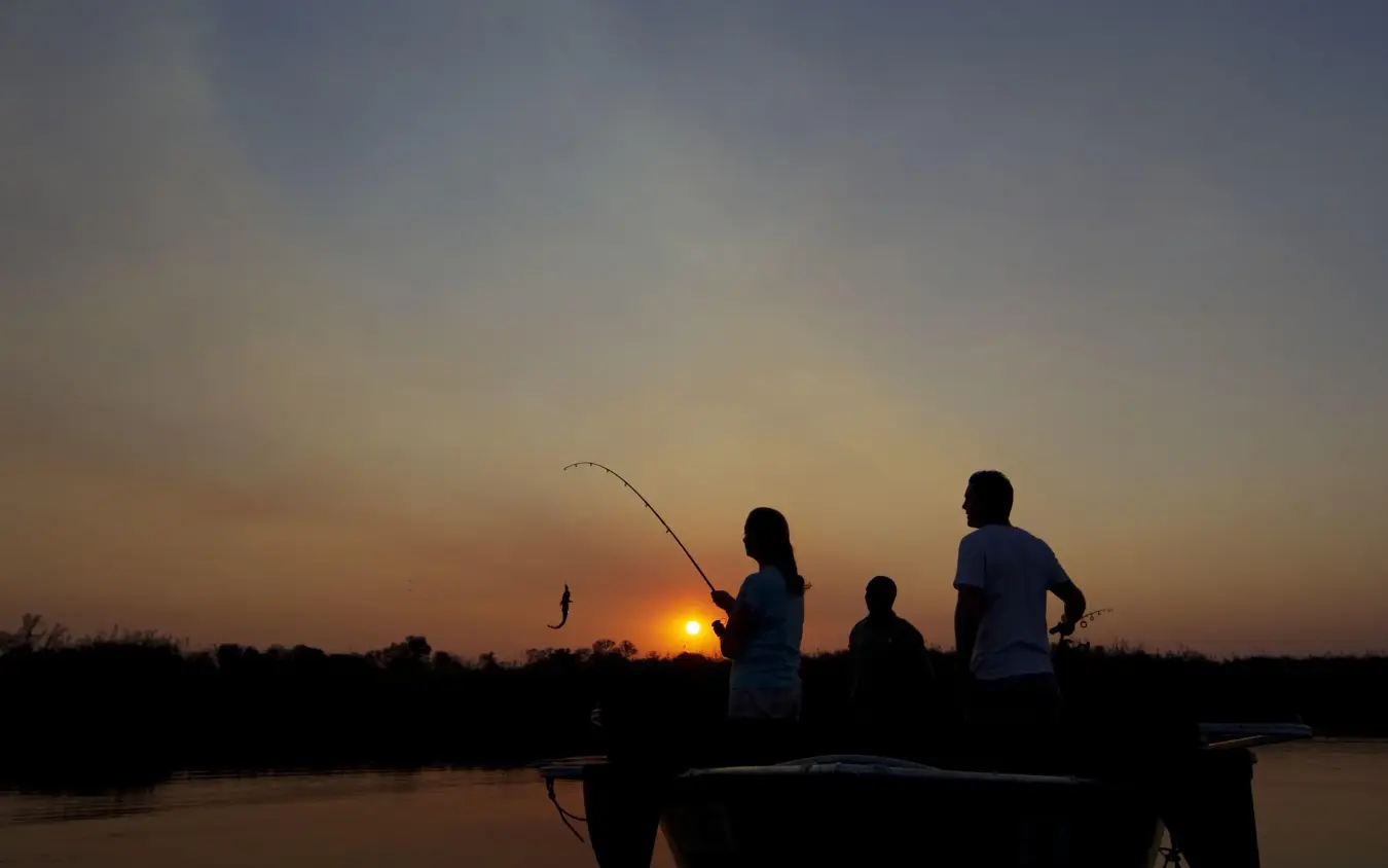 Guests fishing from boat at sunset over water, Kanana, The Okavango Delta, Botswana