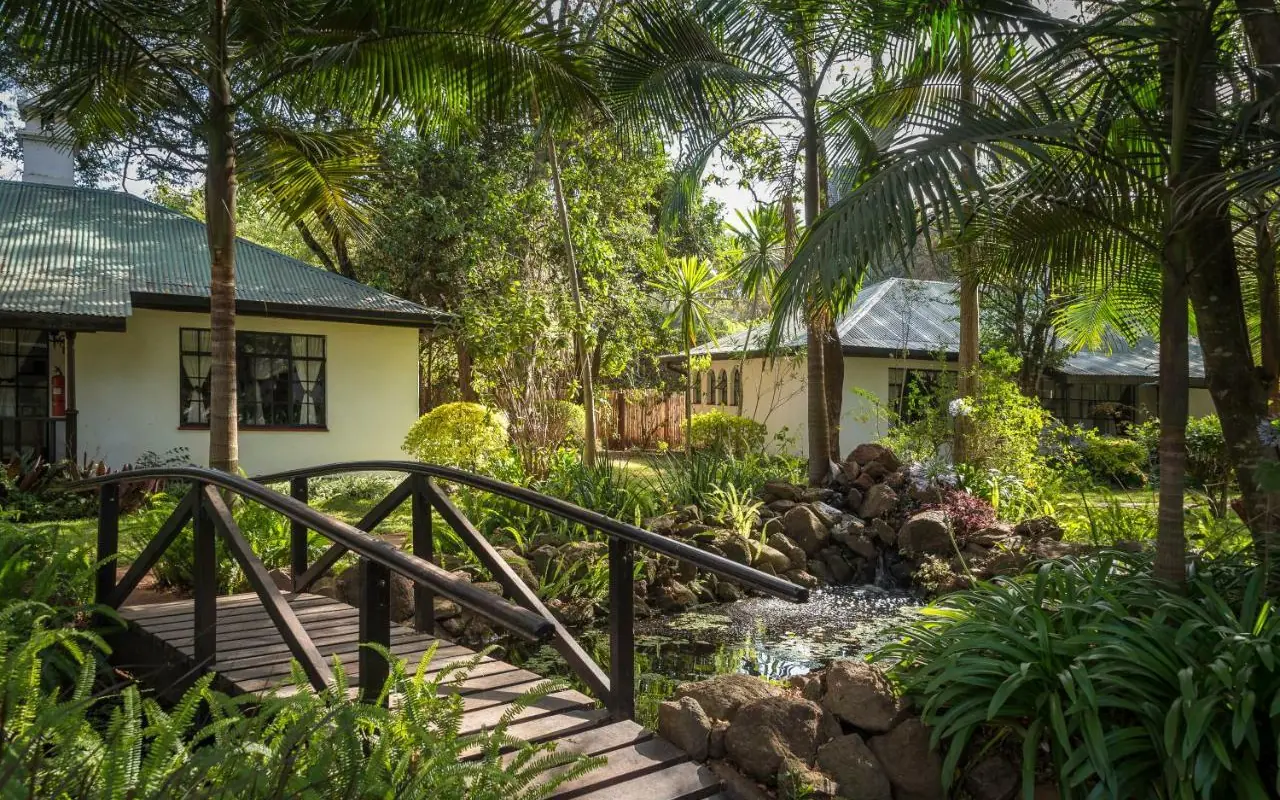 Wooden bridge over stream with tropical bungalows nestled in lush forest garden, Karen Blixen Cottages, Nairobi, Kenya