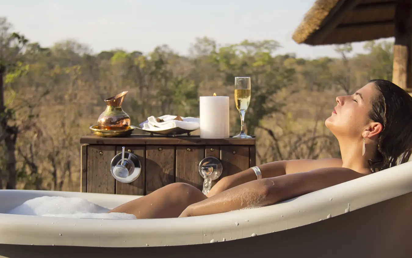 Woman in outdoor bathtub with champagne overlooking forested landscape, Khulu Bush Camp, Hwange National Park, Zimbabwe
