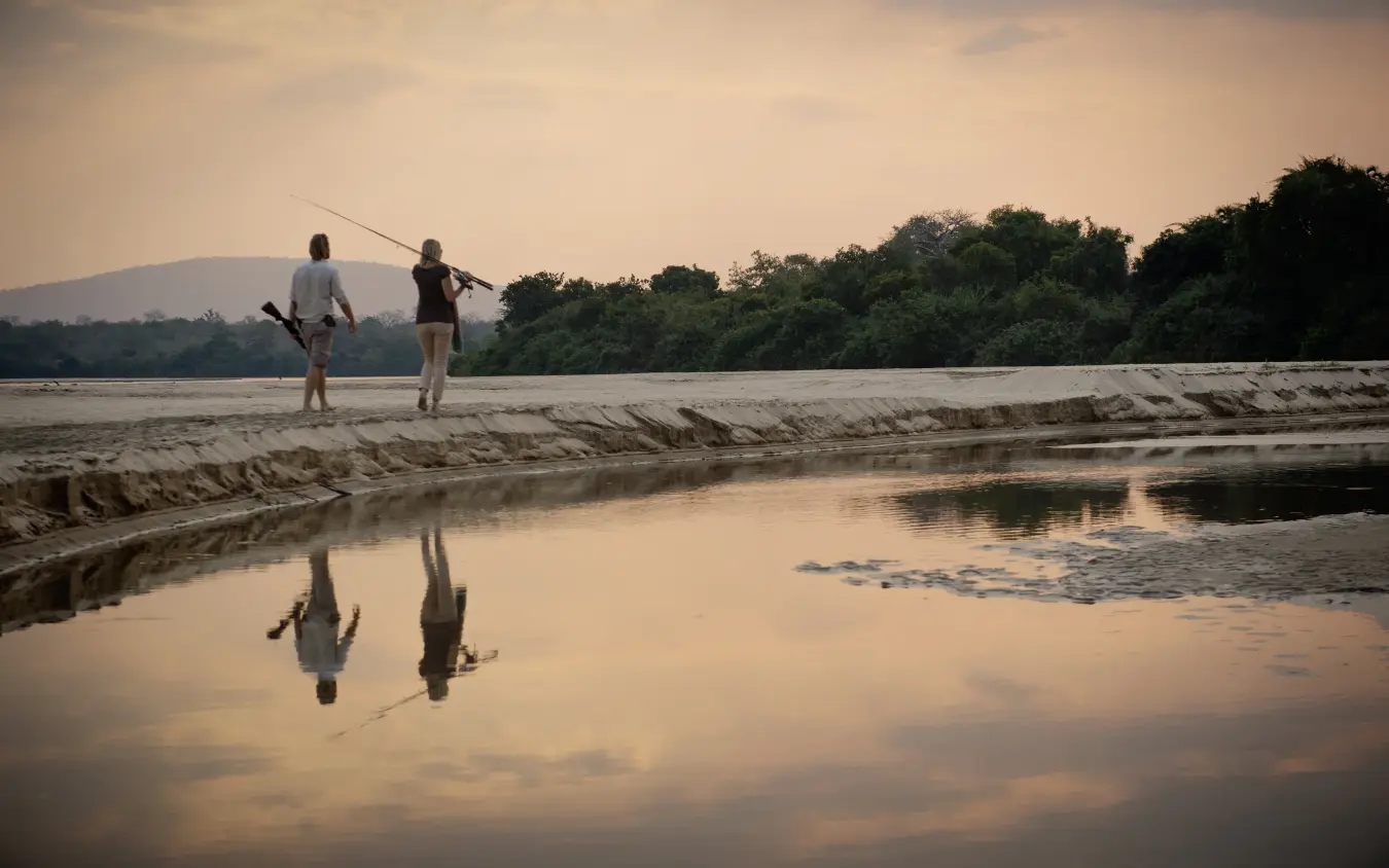Two anglers fishing in shallow river at sunset with forested banks, Kiba Point, Nyerere National Park, Tanzania